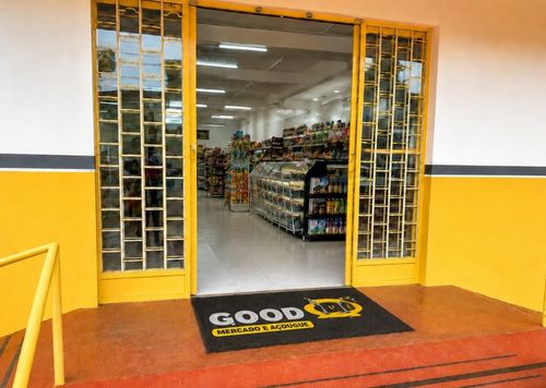 Capacho de vinil preto e amarelo personalizado com a logomarca Good Mercado e Açougue, posicionado na entrada aberta de uma loja com portas de vidro amarelas e prateleiras de produtos ao fundo, piso vermelho, fotografia realista de fachada comercial de alta circulação