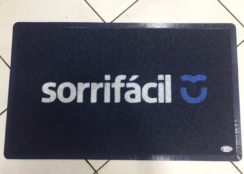 Capacho azul marinho sob medida com a logomarca sorrifácil em branco e ícone de sorriso azul, acabamento com borda de borracha preta sem limite de formato, fotografia realista de tapete personalizado fabricado no tamanho exato