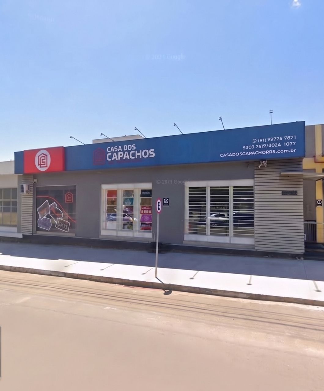 Fachada da loja Casa dos Capachos com letreiro azul e vermelho destacando o nome da empresa, vitrines com produtos e capachos expostos, fachada de revestimento cinza com placas de sinalização, calçada larga e rua em dia ensolarado de céu azul claro, fotografia realista de fachada comercial em Novo Hamburgo